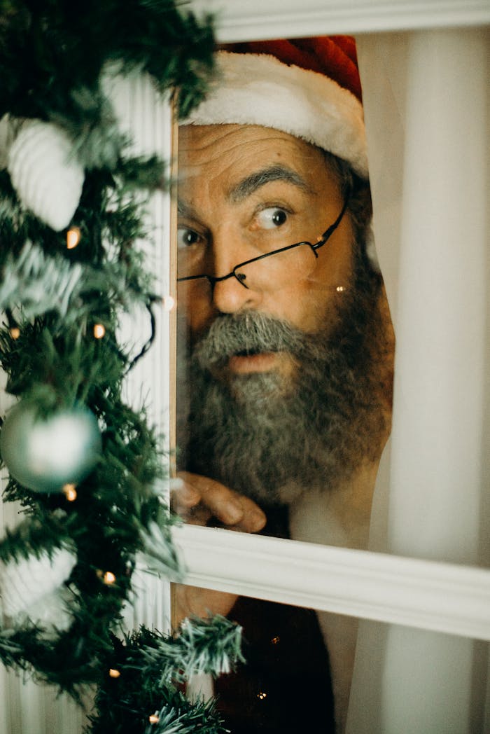 A bearded man dressed as Santa Claus peeking through a decorated window indoors.